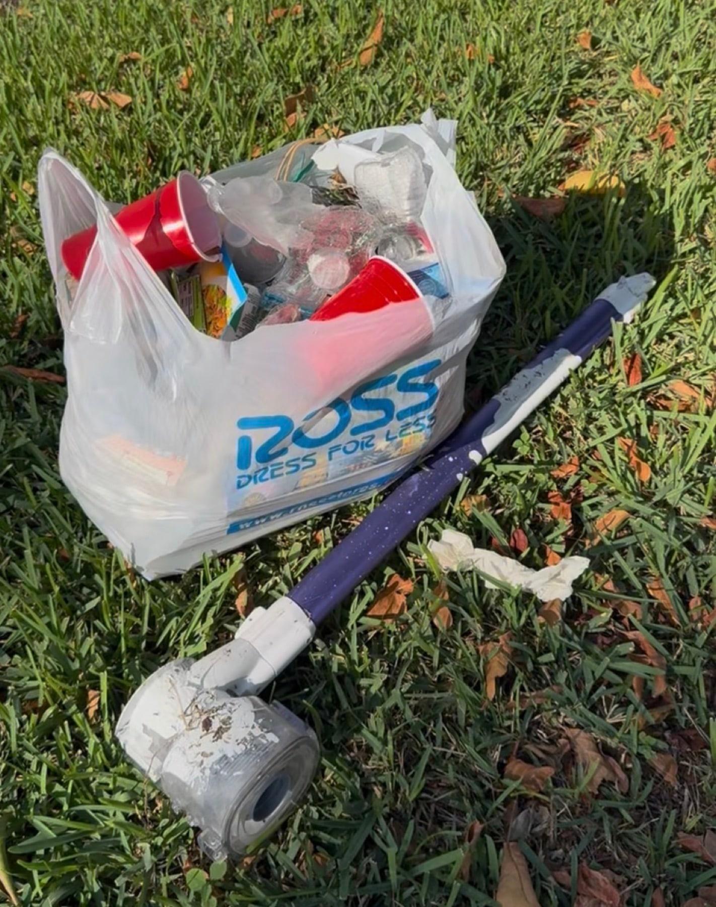 A bag of cups, bottles, and cans next to a grabber tool on the grass